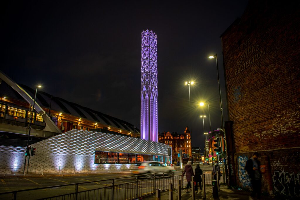 A tall chimney like structure lit up in purple is seen alongside a city centre road.